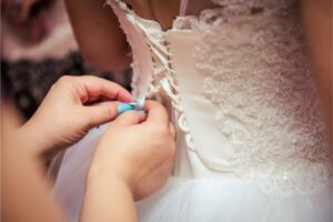 A bridesmaid's hands lacing up the back of a bride's white wedding dress detailed with lace and beads.